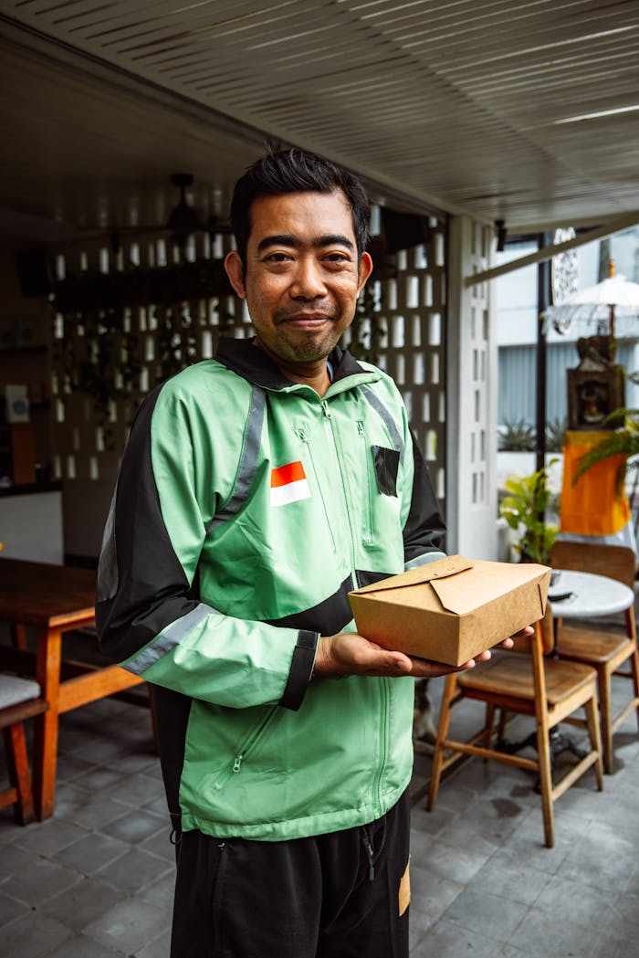 Asian man in a green jacket holding a food delivery box inside a modern café.